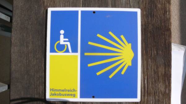 Quelle: Förderverein für den Himmelreich-Jakobusweg e.V. An einem Pfosten befindet sich ein Schild mit der Jakobusmuschel der Pilger*innen, einem Rollstuhlsymbol und dem Schriftzug Himmelreich-Jakobusweg.